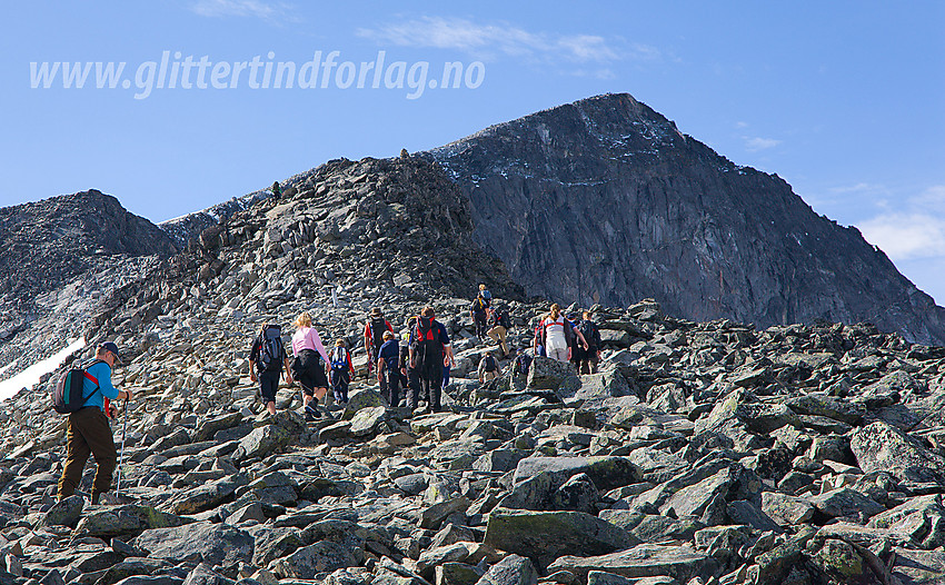 Fjellvandrere på vei mot Galdhøpiggen, som ruver bakenfor. Styggebreen er krysset, og folk tar fatt på fjellryggen som fører til topps.