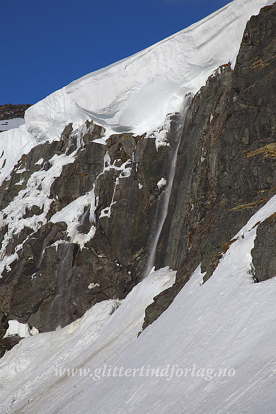 En bekk med smeltevann fra Smørstabbreen kaster seg utfor en fjellvegg ikke så langt fra Krossbu. Over henger en mektig snøskavl.