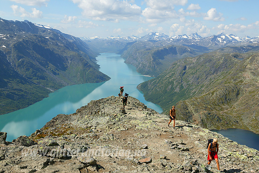 Fjellvandrere på rekke og rad som akkurat har kommet opp den bratteste delen av Besseggen. I bakgrunnen ser man dypt inn i Jotunheimen med Gjende som selve livsnerven.