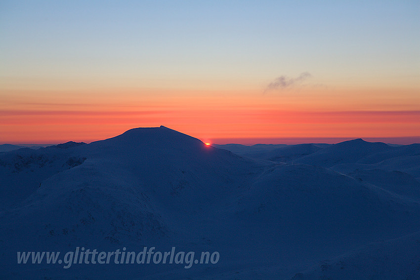 Morgengry en vintermorgen sett fra Galdhøpiggen. Silhuetten til Glittertinden midt i mot. Sola kommer opp like til høyre for denne.