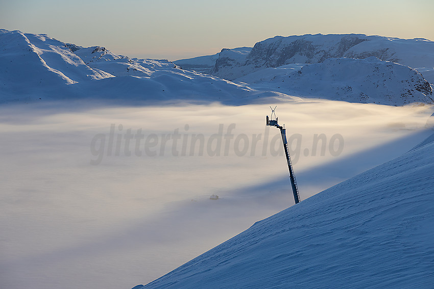 I lia opp mot Galden med tåketeppe over Tyin. Bergsfjellet i bakgrunnen. Installasjonen er en "rasdetonator" som via fjernstyring kan slippe sprengladninger ned på snøfonner under og løse ut snøskavler under kontrollerte former.