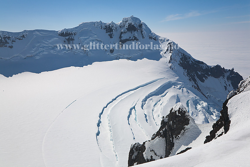 Utsikt fra Hakluyttoppen tvers over krateret til Haakon VII Topp på andre siden. I forgrunnen Sentralkrateret med Weyprechtbreen. Enda lenger i forgrunnen Arnes Pinakkel, en morsom liten sak vendt inn mot krateret. Vi målte toppen til en p.f. på 9.7 meter etter å ha gravd oss ned etter laveste skardpunkt. Toppen kan utmerket ha en p.f. på 10 meter, og høyden er nær 2140 moh.